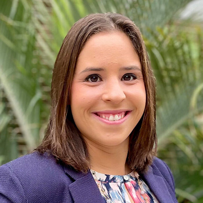 Portrait of a smiling woman with shoulder-length brown hair, wearing a purple blazer, outdoors with green plants in the background.